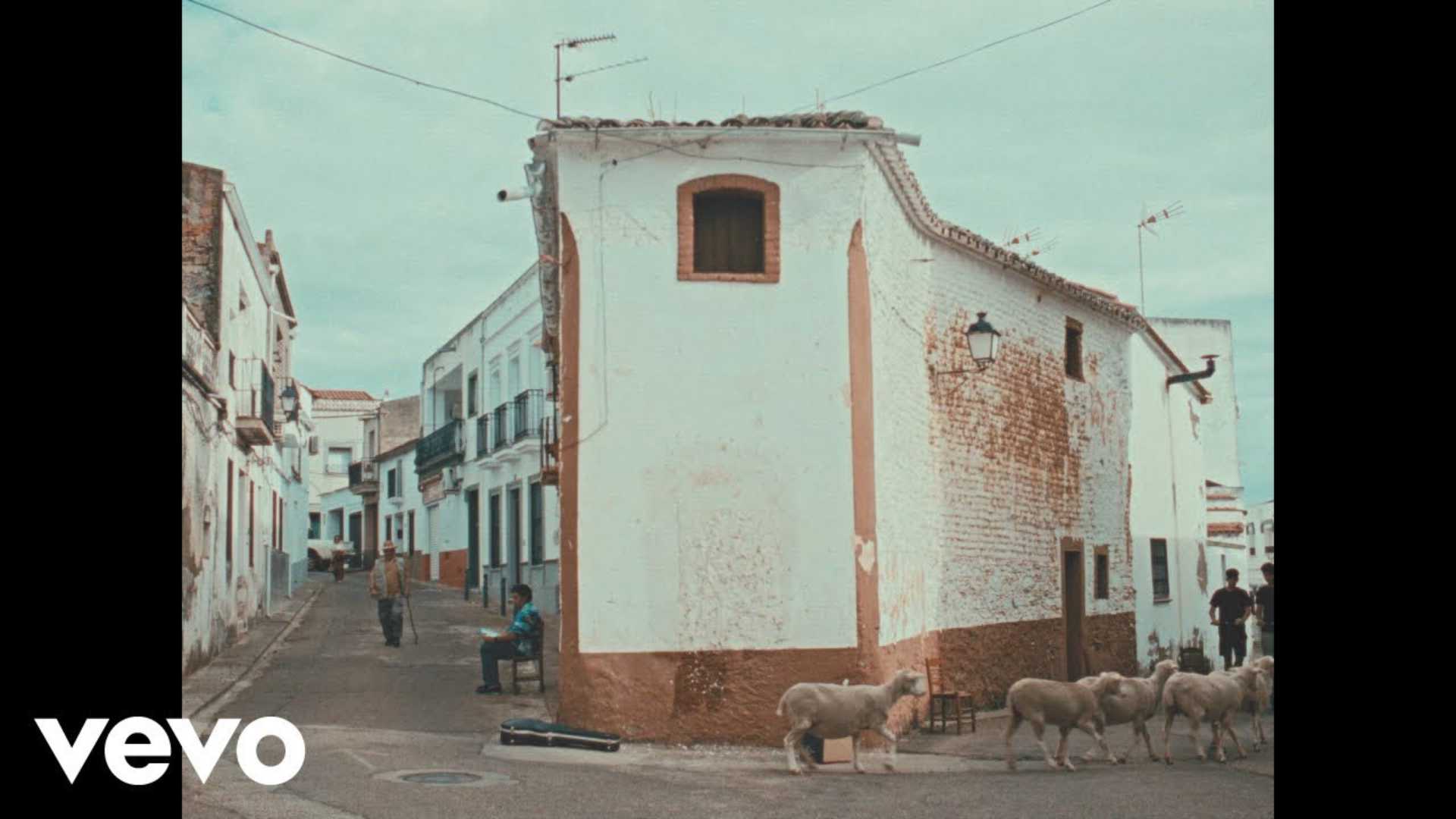 Rebaño de ovejas cruza una calle de un pueblo frente a casas encaladas, con varias personas al fondo y el logotipo de Vevo