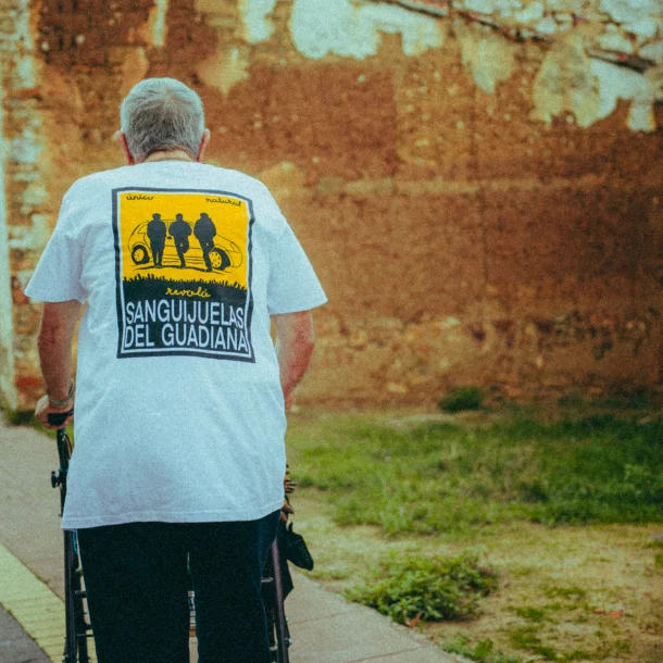 Hombre mayor de espaldas, con camiseta de Sanguijuelas del Guadiana, camina apoyándose en un andador