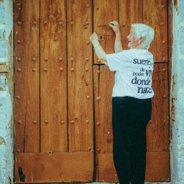 Mujer mayor intenta abrir una gran puerta de madera con remaches, vestida con una camiseta blanca con texto en la espalda