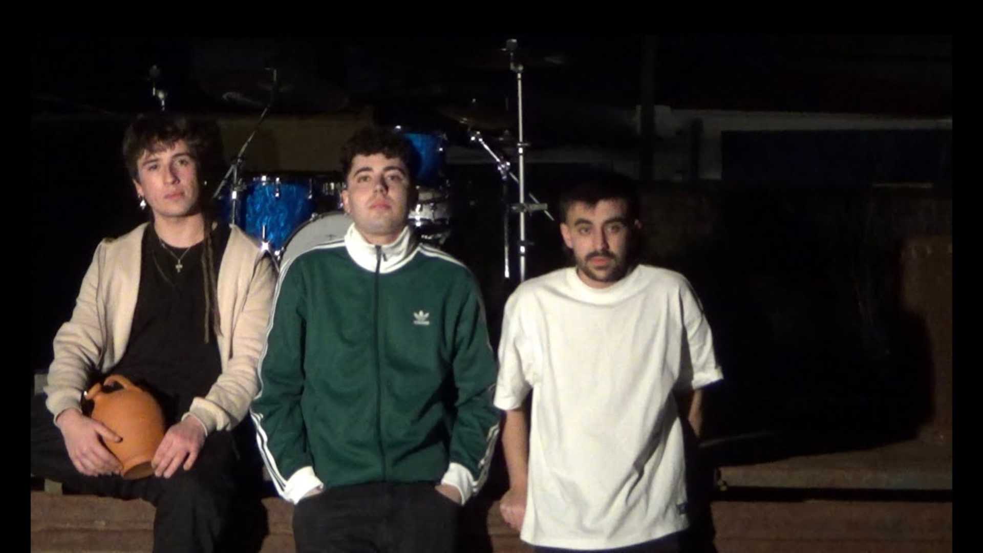 Tres jóvenes sentados frente a una batería en un escenario; uno sostiene un cántaro de barro